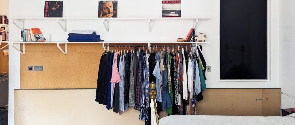 a rack of clothes hanging on a wall with a shelf above holding records