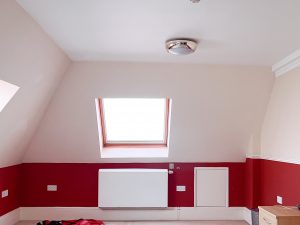 an empty room with white walls and a a strip of red about a metre thick at the bottom of the wall. a white radiator is mounted on the wall and there is an angled ceiling and window in the wall