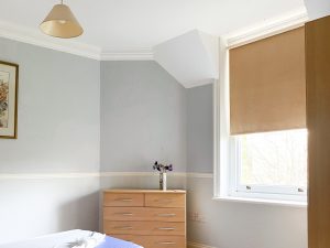 a blue walled bedroom with a window with a brown blind, wooden cabinet with flowers on it, and a framed picture on the wall