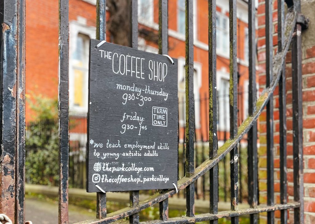 a black sign tied to a fence with an orange brick building in the background. the sign reads 'the coffee shop' and has opening hours, a website link and instagram handle