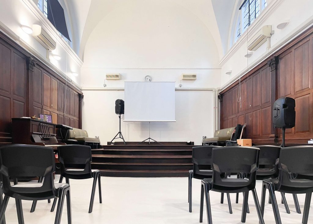 a white-floored hall with dark brown wooden walls and a projector set up. on either side of an aisle sit plastic school chairs