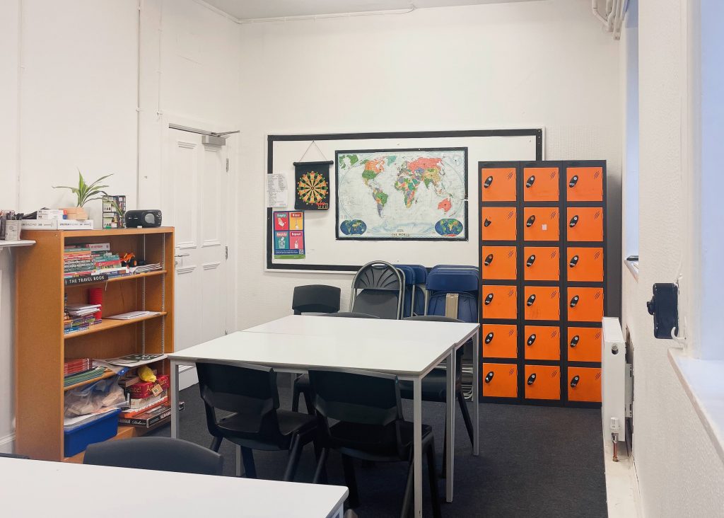 two white desks pushed up against each other in a blue-carpeted floor in a classroom with a world map on the wall and a set of orange and black lockers.