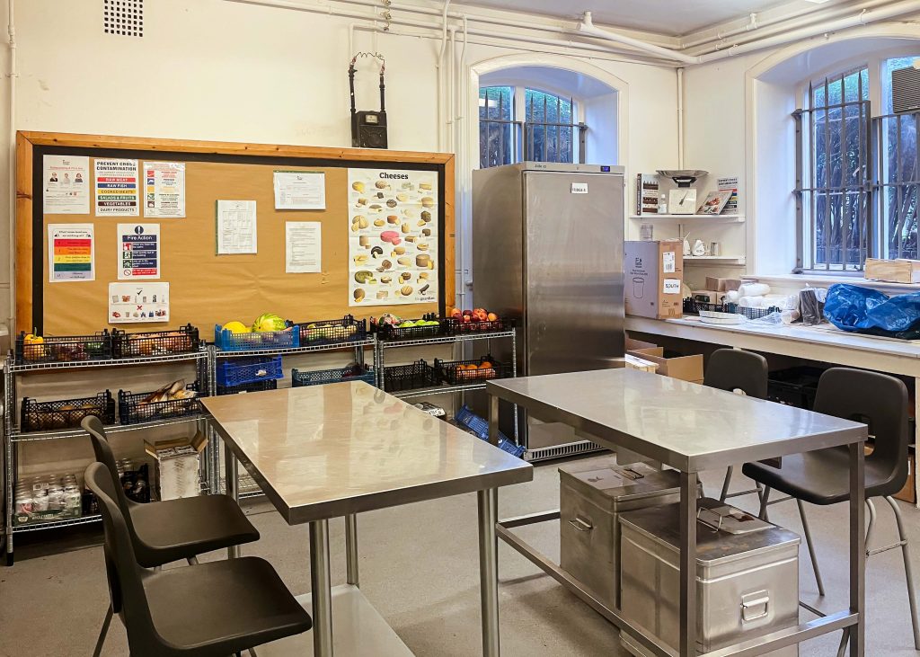 stainless steel tables with black benches sit in a kitchen. a brown wooden-framed board on the wall have posters about food on it and on shelves are baskets of fruit and vegetables