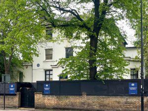 a white building with green trees in front of it and a black fence in front with blue signs on it