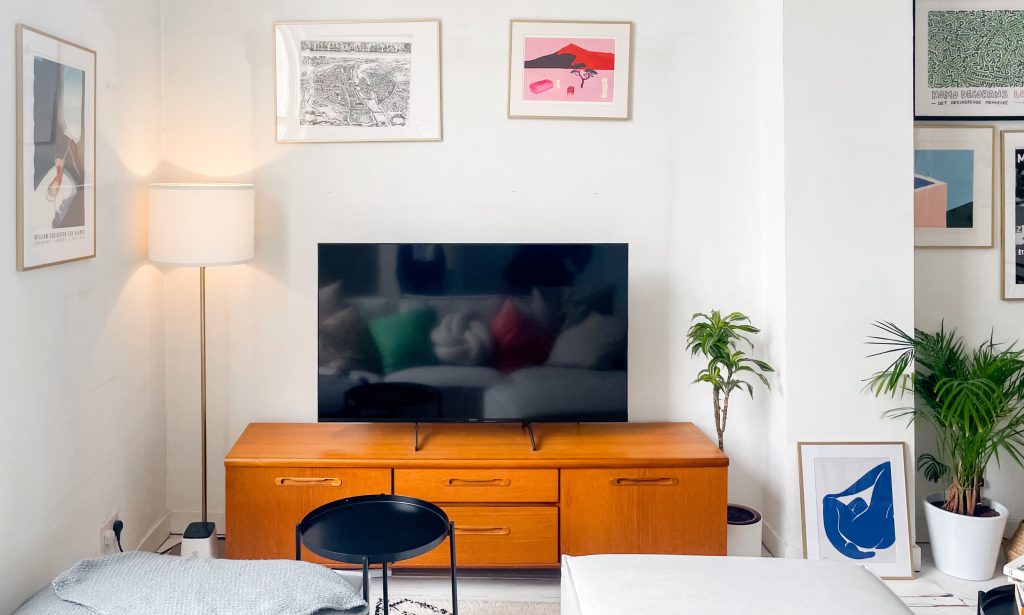 a large tv-screen sits on a brown wooden cabinet with a plant next to it and white framed images on the wall