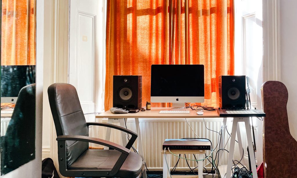 Orange curtained room with a desk setup with a mac desktop and speakers on either side, a black desk chair sits next to the desk