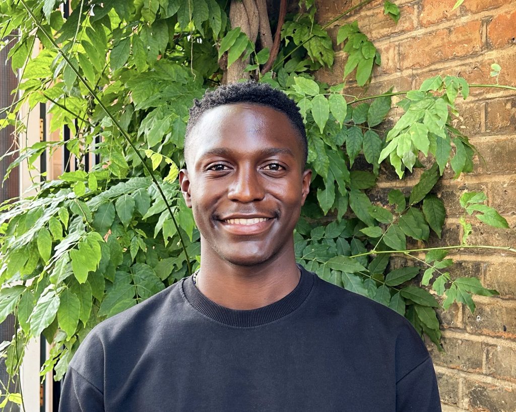 man in black tshirt with a green plant behind him smiles at camera