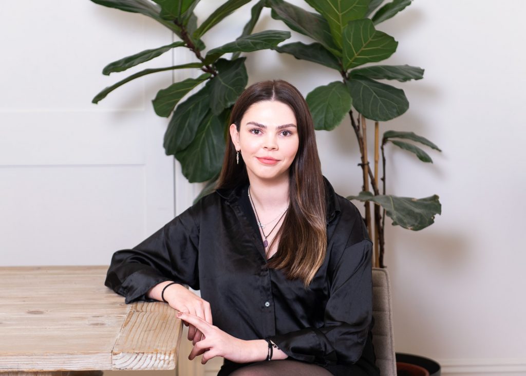a woman with dark hair in a black silk shirt smiles at camera. behind her is a large green plant