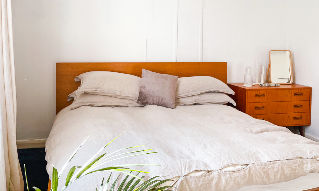 a bed with a wooden frame and grey bedding and a green plant is in front