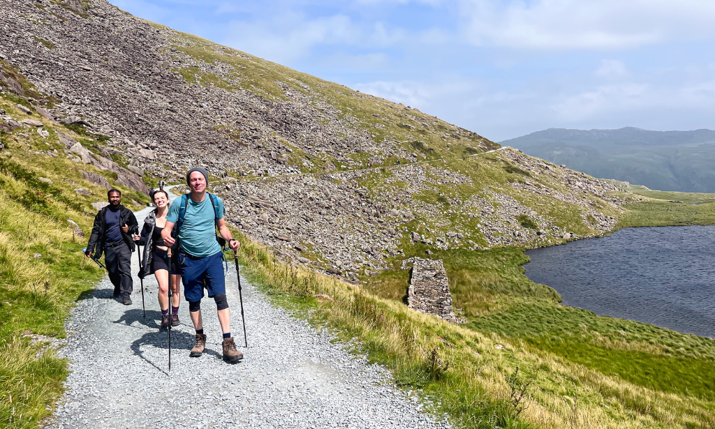 three hikers on a path surrounded by green hills with a lake to their left