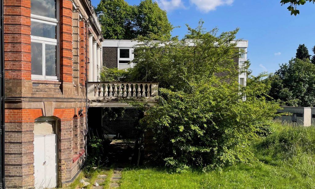 a red brick building with a balconty and overgrown greenery in front
