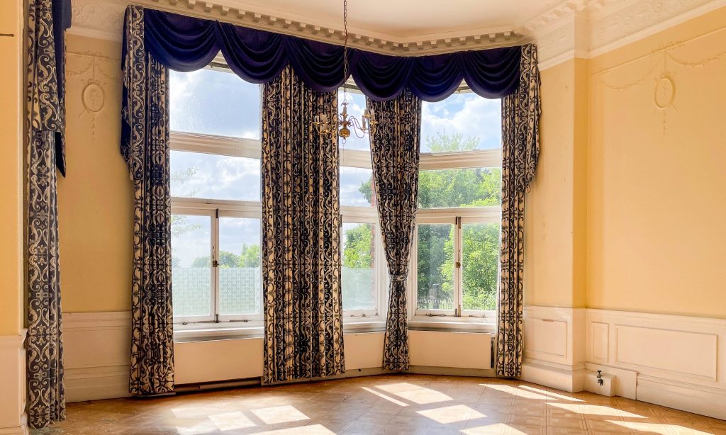 a yellow-walled high-ceiling room with a wooden floor, floor to ceiling windows and blue curtains